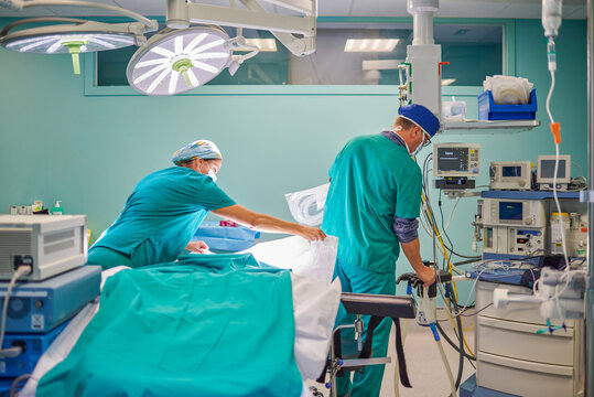 Back view of unrecognizable male and female medical assistants in uniforms preparing couch and devices in operating room for surgery in modern hospital