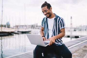 Positive young african american man in eyewear for vision correction typing on laptop computer share content online in personal blog, smiling dark skinned hipster guy enjoying studying on free time