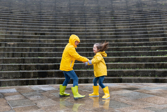 Side view of delighted siblings in yellow raincoats and rubber boots holding hands and having fun on wet street in rainy day while smiling and looking at each other