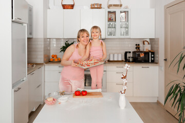 Mom and daughter cook in the kitchen. A young woman cooks with her child in the kitchen at home. Girls in pink clothes cook pizza. Happy and cheerful mom and daughter