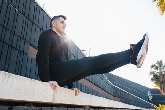 Full length fit strong sportsman in black activewear performing L sit exercise on concrete fence railing during intense work out on sunny street