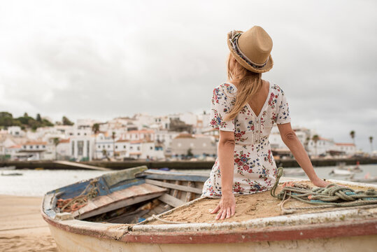 Back view of unrecognizable young woman in elegant dress and sunhat sitting on old shabby wooden boat on sandy beach and admiring river and town architecture during trip in Ferragudo