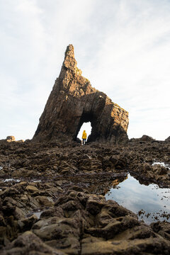 Distant traveler in warm jacket standing on natural arch of rough stiff rock located on stony vast coast on clear autumn day