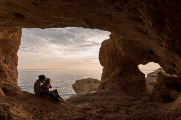 Side view of man and woman sitting in entrance of rocky cave near sea in Algar seco caves in Algarve, Portugal