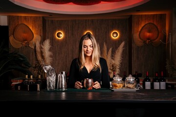 Self assured focused young female barkeeper with long blond hair in stylish outfit preparing cocktail in metal mug standing at counted in stylish bar