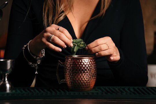 Cropped unrecognizable self assured focused young female barkeeper with long blond hair in stylish outfit decorated cocktail with lemon slices while standing at counted in stylish bar