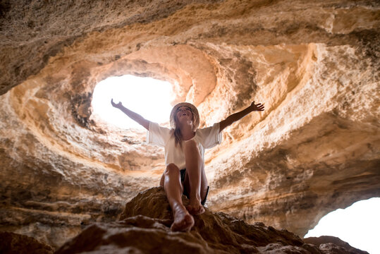From below cheerful female in hat sitting in rock with outstretching arms looking at hole in ceiling of cave near sea on sunny day in Algarve, Portugal