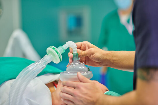 Anonymous male anesthesiologist in medical uniform and cap holding anesthesia mask on face of unrecognizable patient lying on couch in operating room