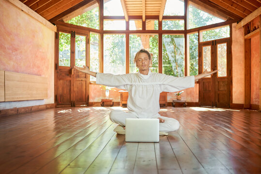 Young concentrated male sitting with closed eyes and crossed legs while practicing yoga against netbook in meditation room