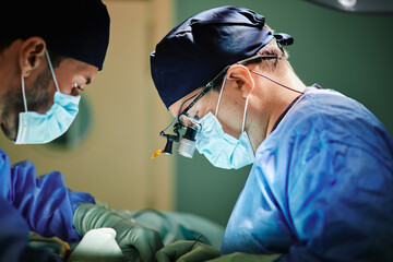 Side view of unrecognizable male doctor with assistant in medical gowns and masks performing surgery with laser in operating room