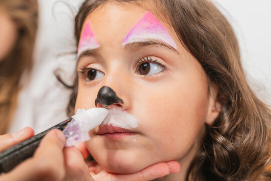 Crop creative artist applying colorful body art paints on cute little girl face in light studio