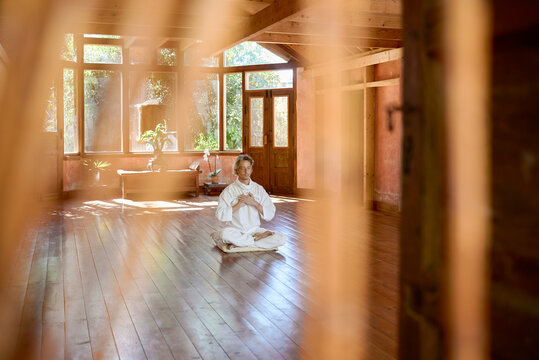 Young mindful male barefoot male sitting in lotus pose on pillow with eyes closed practicing yoga on floor near bowl gong and Buddha statuette