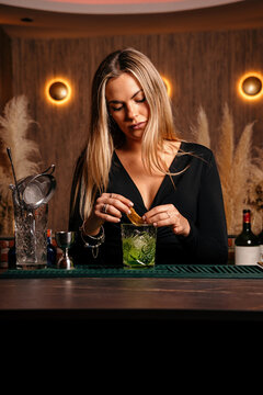 Self assured focused young female barkeeper with long blond hair in stylish outfit decorated cocktail with lemon slices while standing at counted in stylish bar