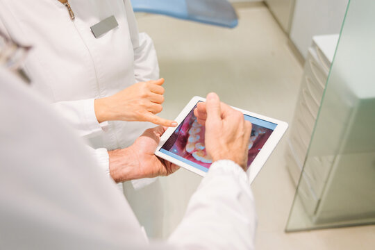From above crop anonymous dentists in medical robes examining teeth condition on x ray scan on tablet while working together in modern dental clinic