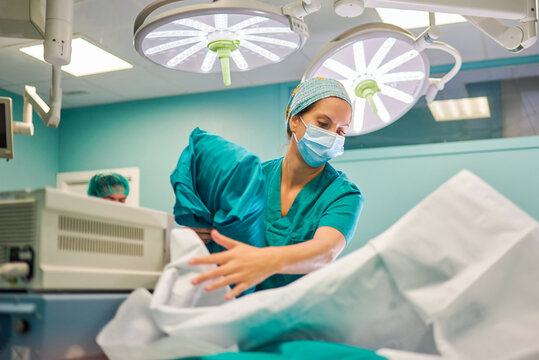 From below female medical assistant in uniform preparing bed couch with clean sheets in operating room for surgery in modern hospital