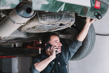 Young focused male auto master speaking on cellphone while checking up rear part of transport and looking up in workshop