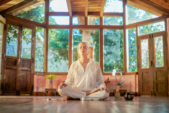 Young Mindful Male Barefoot Male Sitting In Lotus Pose Practicing Yoga On Floor Near Bowl Gong And Buddha Statuette