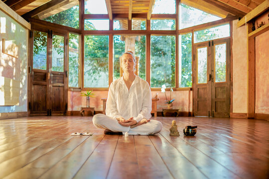 Young mindful male barefoot male sitting in lotus pose practicing yoga on floor near bowl gong and Buddha statuette