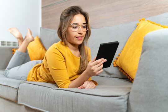 Smiling female lying on soft couch in living room and reading interesting e-book while spending time at home