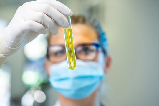 Serious female scientist in mask and latex gloves standing with glass test tube filled with chemical liquid while working in lab