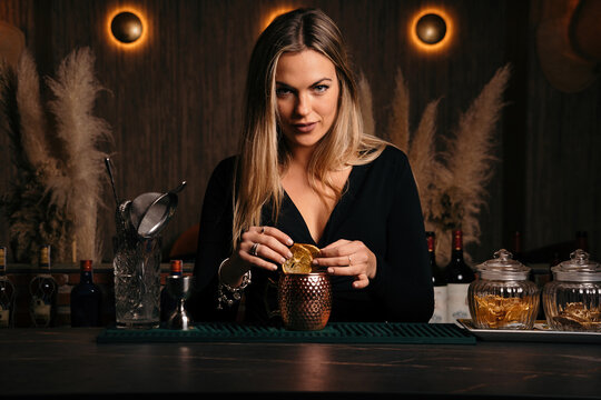 Self assured attractive young female barkeeper with long blond hair in stylish outfit decorated cocktail with lemon slices while standing at counted in stylish bar