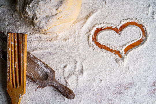 From above of wooden table with heart in wheat flour and bunch of dough with wooden pasta board