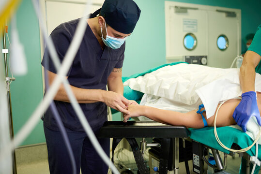 Anonymous young male doctor in medical uniform and mask inserting intravenous catheter in hand of crop faceless patient lying on couch before surgery in operating room
