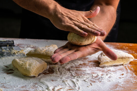 High angle of faceless baker kneading and shaping balls of dough making bread at wooden table in flour