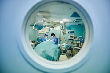 Through round window of unrecognizable doctors in uniforms and masks performing surgery in modern operating room