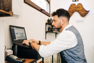 Side view of focused male barber working on netbook with open program on screen in beauty salon near cash register till and card reader