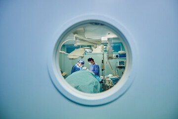 Through round window of unrecognizable doctors in uniforms and masks performing surgery in modern operating room