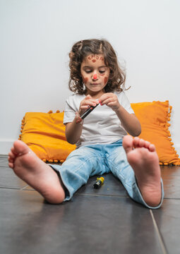 Unemotional little girl in casual wear with red lipstick drawings on happy face while sitting on floor at home