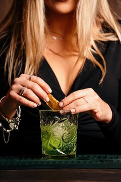 Cropped unrecognizable self assured focused young female barkeeper with long blond hair in stylish outfit decorated cocktail with lemon slices while standing at counted in stylish bar