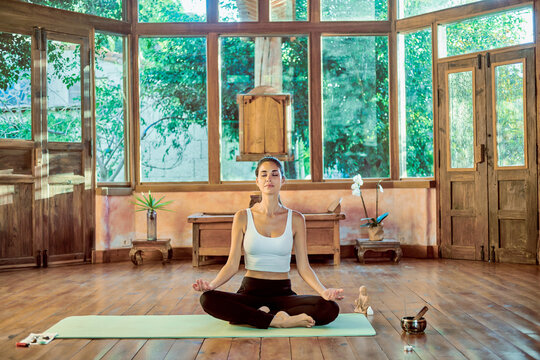 Young concentrated female with closed eyes practicing yoga with crossed legs near bowl gong in house