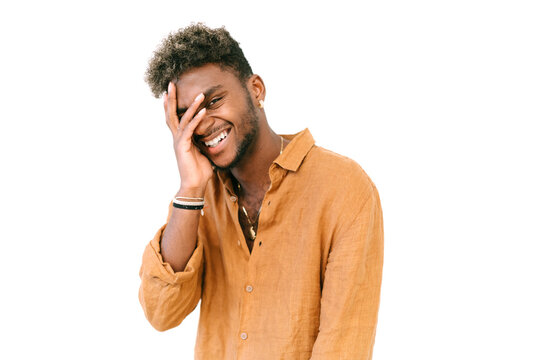 Handsome shy or ashamed young African American guy in casual wear covering face with hand and looking at camera with smile against white background