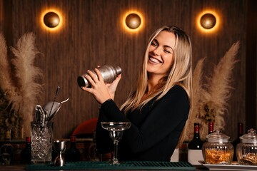 Cheerful young female barkeeper with long blond hair in stylish clothes smiling while mixing cocktail in shaker in modern bar
