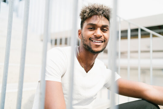 Handsome young bearded hipster black guy with curly hair dressed in casual white shirt looking at camera and smiling while sitting on stairs near metal fence