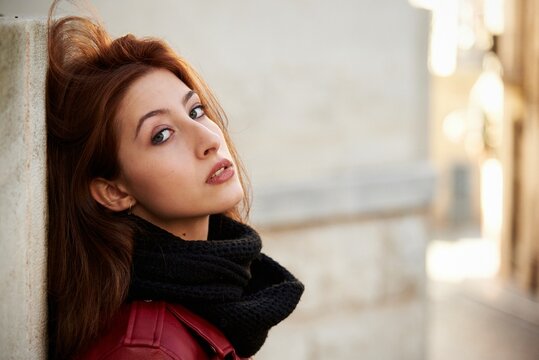 Side view of self assured young lady with long brown hair in trendy red leather jacket leaning on wall of aged shabby building while chilling on street looking at camera