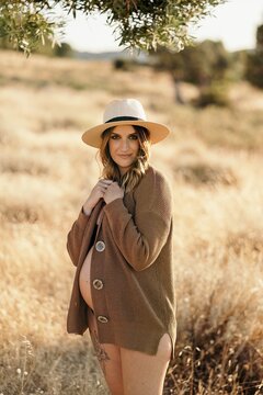 Cheerful pregnant woman wearing a hat lingerie and cardigan standing among dry grass in field placed in countryside and looking at camera in sunny day