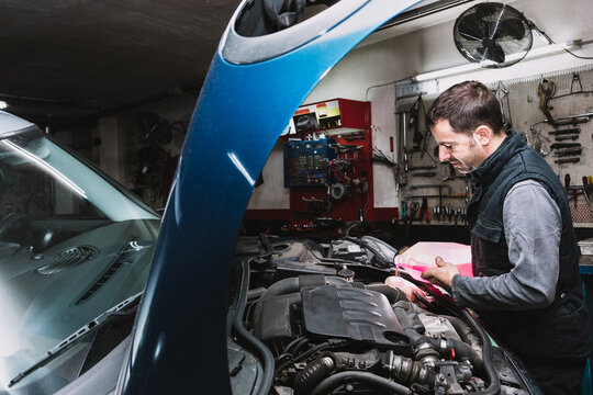 Side view of unshaven maintenance male pouring liquid from canister into radiator in open hood of vehicle in garage