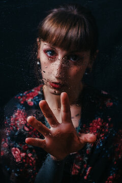 Young sad female in ornamental wear with bang looking at camera behind transparent glass with water drips