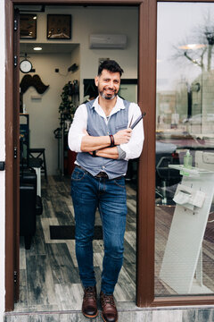Happy smiling male hairdresser in smart casual apparel with professional grooming tools at entrance door of beauty salon in town