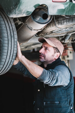 Young attentive bearded auto master in cap revising transport wheel in workshop in sunlight