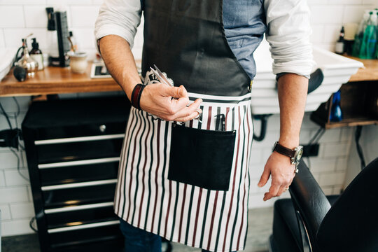 Crop unrecognizable male barber with scissors and comb in striped apron in hairdressing salon