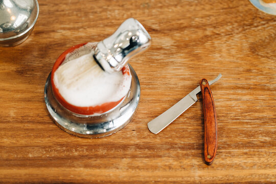 From above of shaving brush with soft bristles in bowl with foamy soap near straight razor on wooden table