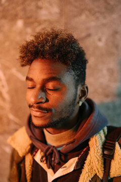 Young African American hipster male with curly hair and beard wearing warm jacket looking at camera while standing against shabby wall