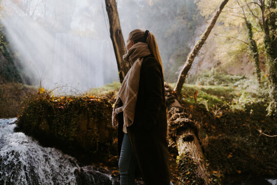 Side view of anonymous female in warm coat standing in forest in autumn and enjoying view of waterfall
