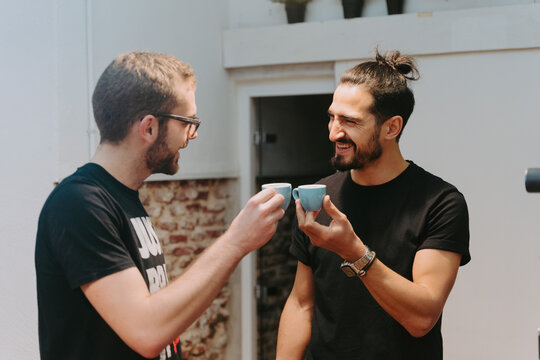 Delighted male baristas with small cups of espresso tasting prepared coffee while clinking mugs and looking at each other