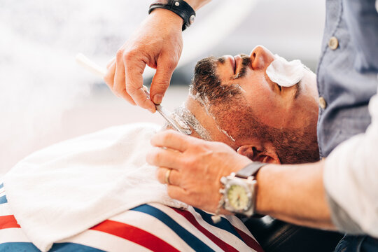 From above of crop anonymous beauty master in wristwatch shaving beard of client with straight razor during steam vapor treatment in hairdressing salon