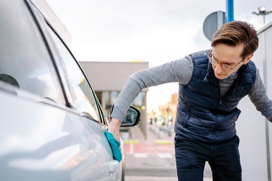 Focused young male in casual clothes and eyeglasses wiping vehicle with rag while standing in car wash station against cloudy sky